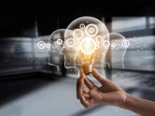 Hand holding glowing light bulb overlayed with illustrated interlocking gears and the outline of a human head.